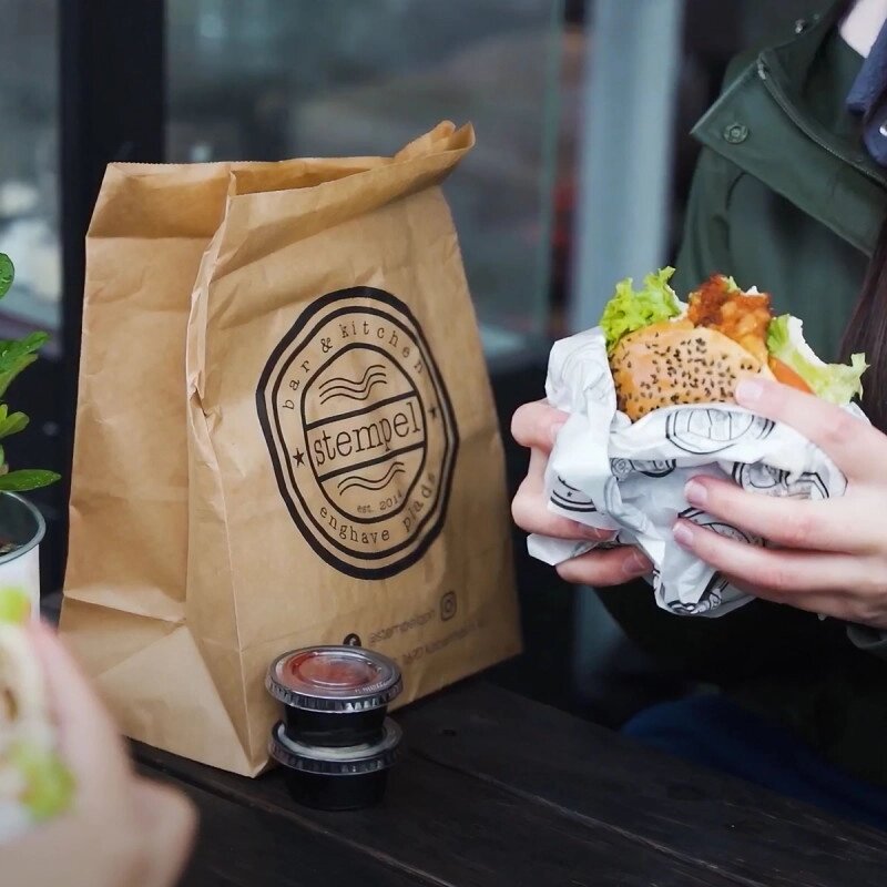 Person enjoying a burger next to a branded paper bag with logo from Stempel, showcasing custom print on takeaway paper bags.